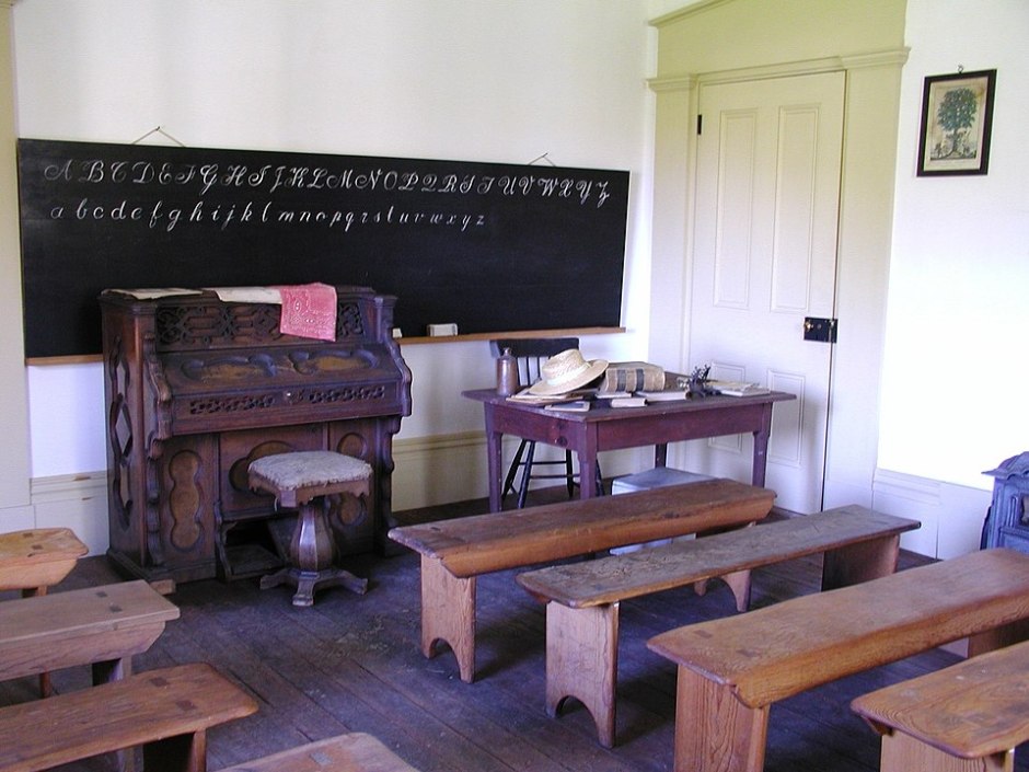 A Restoration-era classroom. A pieano is at the front. Student benches are on the main floor. There's a teacher's desk next to the piano, and behind the piano an old chalkboard with the English alphabet on it.